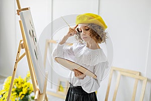 Portrait of cute thoughtful little girl artist in yellow beret who paints picture in studio while holds color pallete