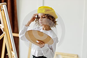 Portrait of cute thoughtful little girl artist in yellow beret who paints picture in studio while holds color pallete