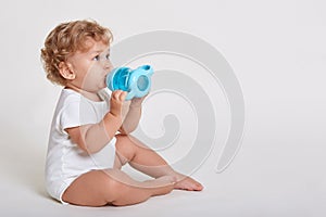 Portrait of cute toddler drinking water from bottle while sitting against white wall, wearing body suit, one year old kid with