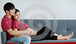 Portrait of cute young Asian couple husband and wife sitting together on a couch at home. A man smiling and pointing something