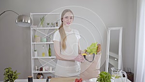 Portrait of cute young Caucasian girl posing with vegetables bag. Beautiful woman standing in the kitchen with healthy