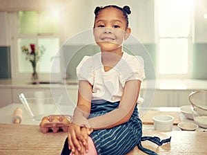 Portrait of a cute young mixed race girl sitting on the kitchen counter smiling and wearing an apron looking thoughtful