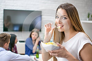 Portrait of cute young woman eating potato chips while sitting with her friends at home.