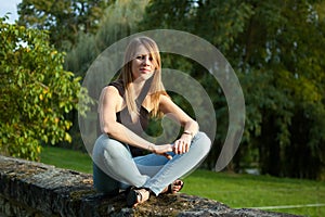 Portrait of the Cute Young Woman Sitting in the Park During Sunset in Jeans and Black Shirt.