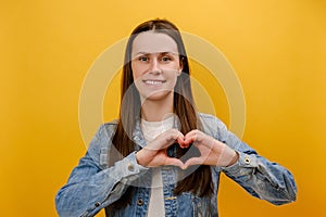 Portrait of cute young woman standing with love gesture, smiling looking at camera, showing heart shape gesture, wearing denim