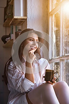 Portrait cute young woman in white shirt drinking tea and looking through window while sitting at windowsill indoors