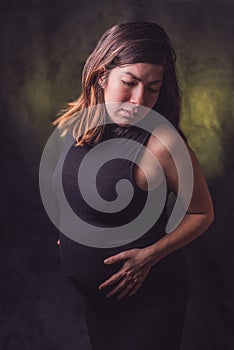 Dark-haired pregnant woman in a black tight dress holding her big gestation belly with her hands