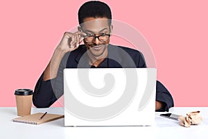 Portrait of dark skinned man works with laptop. Handsome black male sits at desk wears suit, touches his spectacles with hand,