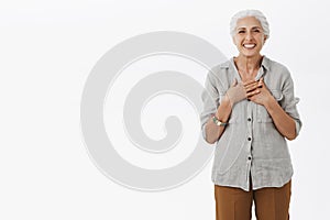 Portrait of delighted charming old female with grey combed hair holding palms on chest with pleased and grateful smile