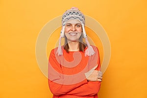 Portrait of delighted confident woman wearing orange sweater and knitted earflap hat, crossed her arms, looking at camera with