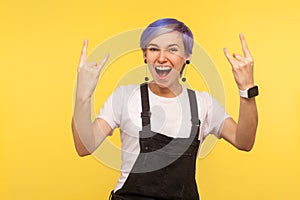 Portrait of delighted funky hipster woman showing rock and roll gesture, crazy punk sign. yellow background, studio shot