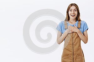 Portrait of delighted greatful charming kind young girl in brown overalls holding palms pressed to chest in thankful