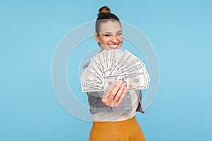 Portrait of delighted happy woman with hair bun in modern outfit holding fan of dollars to camera and smiling