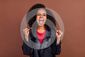 Portrait of delighted pretty carefree girl raise fists scream yes isolated on brown color background