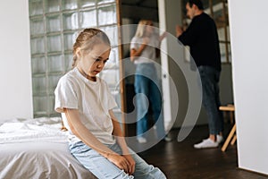 Portrait of depressed cute little daughter sadness looking down sitting on couch during parents quarrelling and fighting