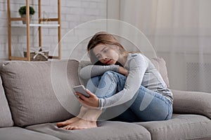 Portrait of depressed young woman sitting on couch with smartphone in her hands