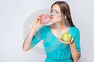 Portrait of desiring young beautiful girl in blue blouse standing, holding pink donut and green apple in the hands and wanted photo