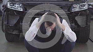 Portrait of desperate Caucasian man sitting in front of broken car and holding head with hands. Upset adult businessman