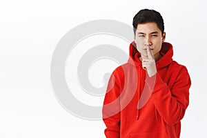 Portrait of devious good-looking asian man in red hoodie, keep secret, squinting mysterious and shush, make hush gesture