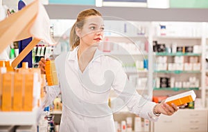 Portrait of diligent smiling pleasant positive female druggist in white coat working in pharmacy
