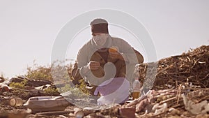 Portrait of a dirty homeless hungry man in a dump eating orange for food in the package with walking goes looking for