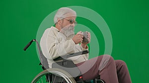 Portrait of disabled man isolated on chroma key green screen close up. Senior man sitting in wheelchair holding cup of