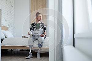 Portrait of disabled young man reading newspapers on bed indoors at home, leg prosthetic concept.