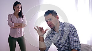 Portrait of disappointed guy after quarrel with female sitting at home folding hands near face and woman standing