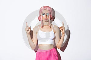 Portrait of disappointed silly girl in pink wig, pointing fingers up and sulking upset, complaining about something bad