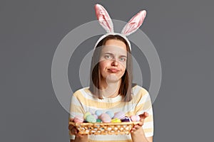Portrait of disappointed woman wearing rabbit ears holding colorful Easter eggs isolated on gray background, looking at camera
