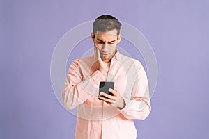 Portrait of disappointedyoung man holding phone with doubtful and skeptical expression on pink isolated background.