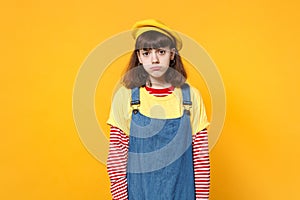 Portrait of disconcerted upset girl teenager in french beret and denim sundress standing isolated on yellow wall