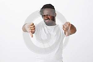 Portrait of disgusted or displeased african-american businessman in white t-shirt, showing thumbs down, frowning and