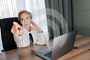 Portrait of displeased middle-aged business woman boss sitting in office desk, pointing finger at someone, looking at