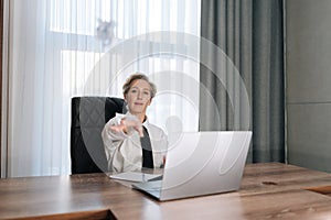 Portrait of displeased middle-aged female boss sitting in office desk, gesturing with hand at someone, looking at camera