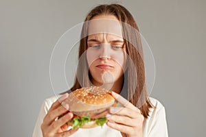 Portrait of dissatisfied sad woman wearing white T-shirt holding burger isolated on gray background, thinking eat or not, proper