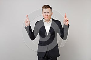 Portrait of dissatisfied young business man in classic black suit, shirt keeping fingers crossed isolated on grey