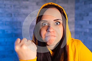 Portrait of dissatisfied young woman showing fist. Close up of distraught female in yellow hood shows fist, looking at