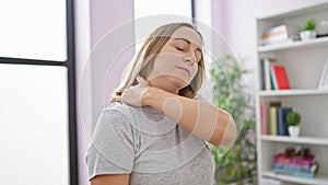 Portrait of a distressed, attractive, young blonde woman suffering from a severe neck ache, standing unhappily indoors within her