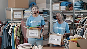 Portrait of the diverse volunteers holding cupboard boxes with clothes and looking at the camera with smile. Shelves