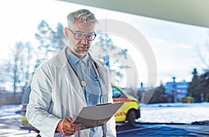 Portrait of doctor in hospital corridor. Handsome doctor with gray hair wearing white coat, stethoscope around neck