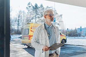 Portrait of doctor in hospital corridor. Handsome doctor with gray hair wearing white coat, stethoscope around neck