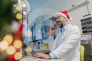 Portrait of doctor phone calling in emergency room, decorated for Christmas. Mature male doctor with Santa hat
