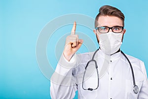 Portrait of doctor wearing a mask and white uniform pointing up with a finger on blue background with copy space