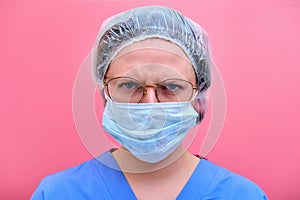 Portrait of a doctor woman with angry facial expression, close up. Nurse in a blue uniform and a medical mask on a pink background
