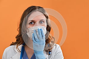 Portrait of a doctor woman with a fright in the eyes, closeup. Nurse in a blue uniform and a protective mask on a orange