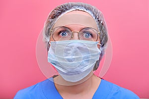 Portrait of a doctor woman with a frightened facial expression, closeup. Nurse in a blue uniform and a medical mask on a pink