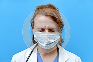 Portrait of a doctor woman with sad facial expression, close up. Nurse in a blue uniform and a medical mask on a blue background