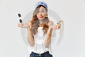 Portrait of doubtful beautiful woman 20s hesitating while holding two pairs of sunglasses, isolated over white background