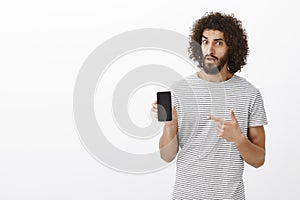 Portrait of doubtful disbelieving attractive male with curly hair, holding smartphone and pointing with index finger at